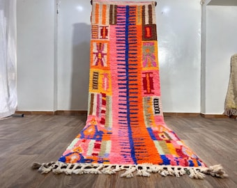 May include: A vibrant, rectangular rug with a colorful geometric design. The rug features a central vertical blue stripe, surrounded by orange, pink, and brown blocks. The bottom edge has a white fringe. The rug is displayed on a wooden floor.