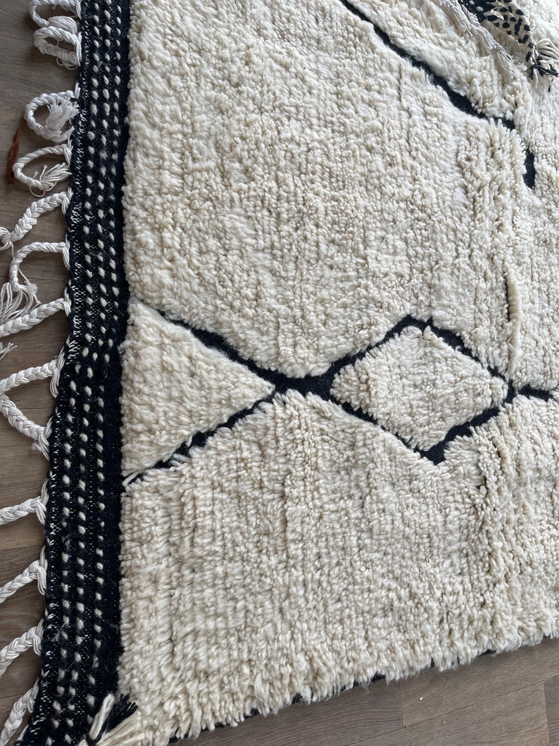May include: A white and black Moroccan rug with a geometric pattern. The rug has a fringe border and is made of wool.
