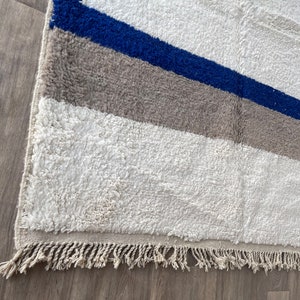 May include: A white rug with blue and gray geometric patterns. The rug has a fringe border.