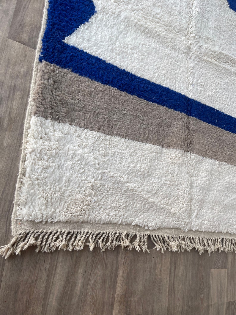 May include: A white rug with blue and gray geometric patterns. The rug has a fringe border.