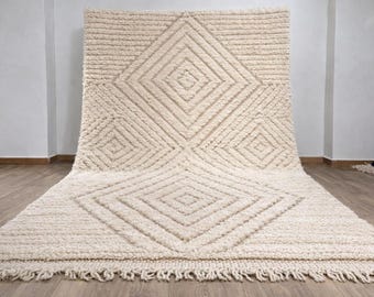 May include: A large, off-white rug with a geometric diamond pattern. The rug has a thick, textured pile and a fringe along the bottom edge. The design features a series of diamond shapes, creating a visually appealing texture.