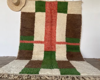 May include: A geometric patterned rug with a white background and brown, green, and pink squares. The rug has a fringe border.
