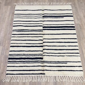 May include: A rectangular area rug featuring an off-white base with horizontal black stripes of varying widths. The rug has a textured pile surface and is finished with a fringe of off-white tassels along the top and bottom edges.