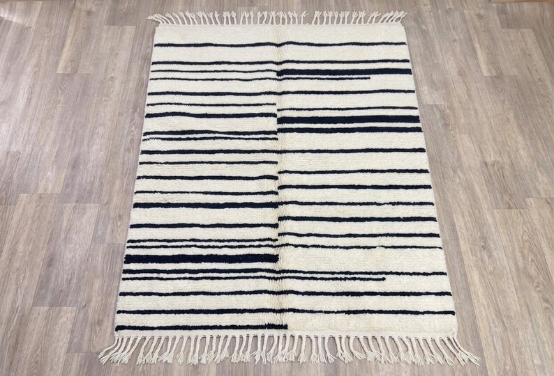 May include: A rectangular area rug featuring an off-white base with horizontal black stripes of varying widths. The rug has a textured pile surface and is finished with a fringe of off-white tassels along the top and bottom edges.