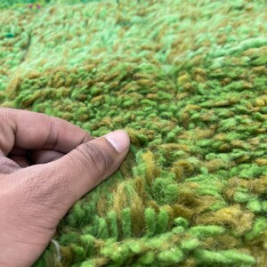 Pu&ograve; includere: Primo piano di un tappeto shaggy verde e marrone con una mano che tocca la trama. Il tappeto presenta un gradiente di colori verde e marrone.