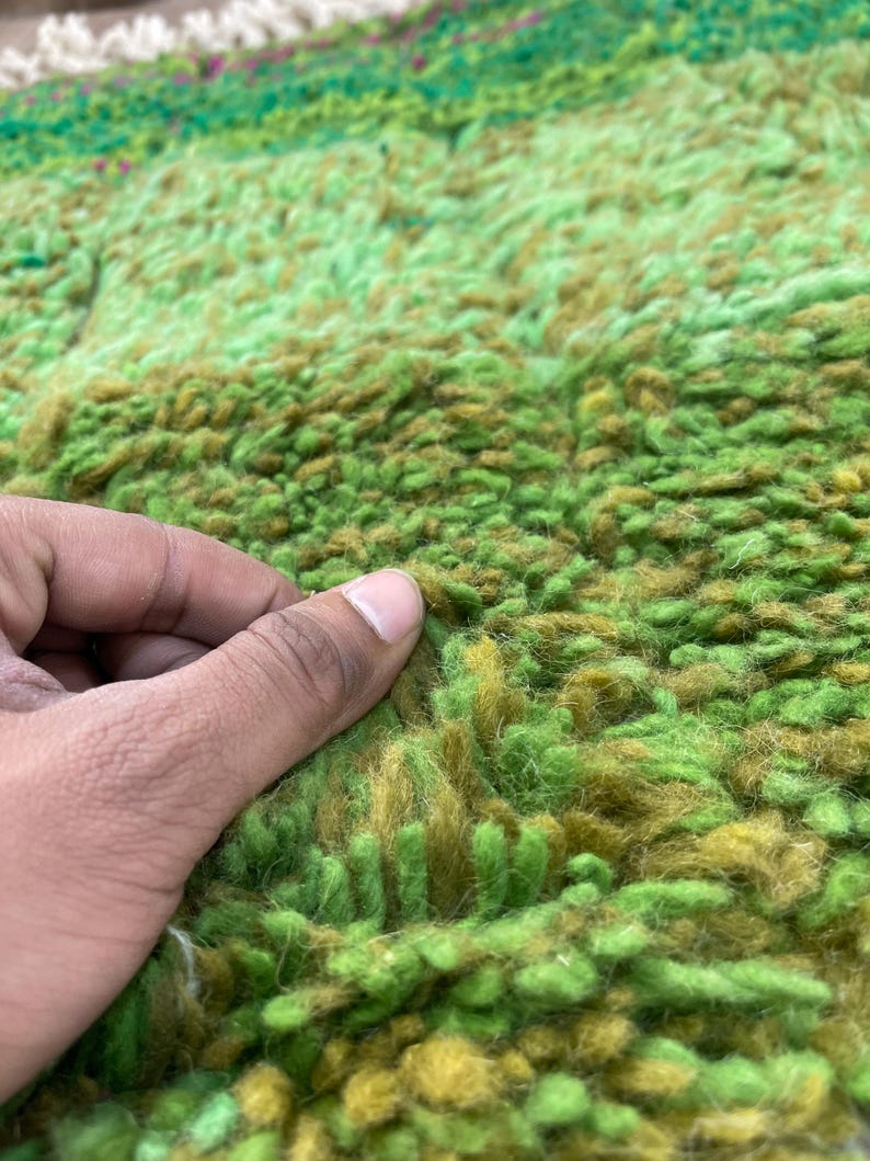 Pu&ograve; includere: Primo piano di un tappeto shaggy verde e marrone con una mano che tocca la trama. Il tappeto presenta un gradiente di colori verde e marrone.