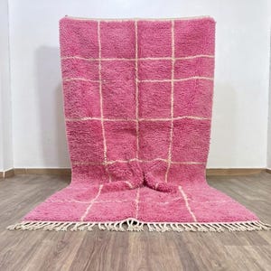 Peut inclure: Un tapis rectangulaire à poils longs d'un rose vif, orné d'un motif à carreaux blanc contrastant. Ce tapis présente une surface épaisse et texturée, terminée par une rangée de franges blanches sur le bord inférieur, formant un motif géométrique.
