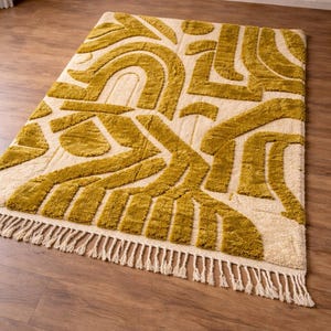 May include: Mustard yellow and cream-colored area rug with an abstract geometric pattern. The rug features a repeating design of curved and angular shapes. The rug has a fringe along one edge. The rug is placed on a hardwood floor.