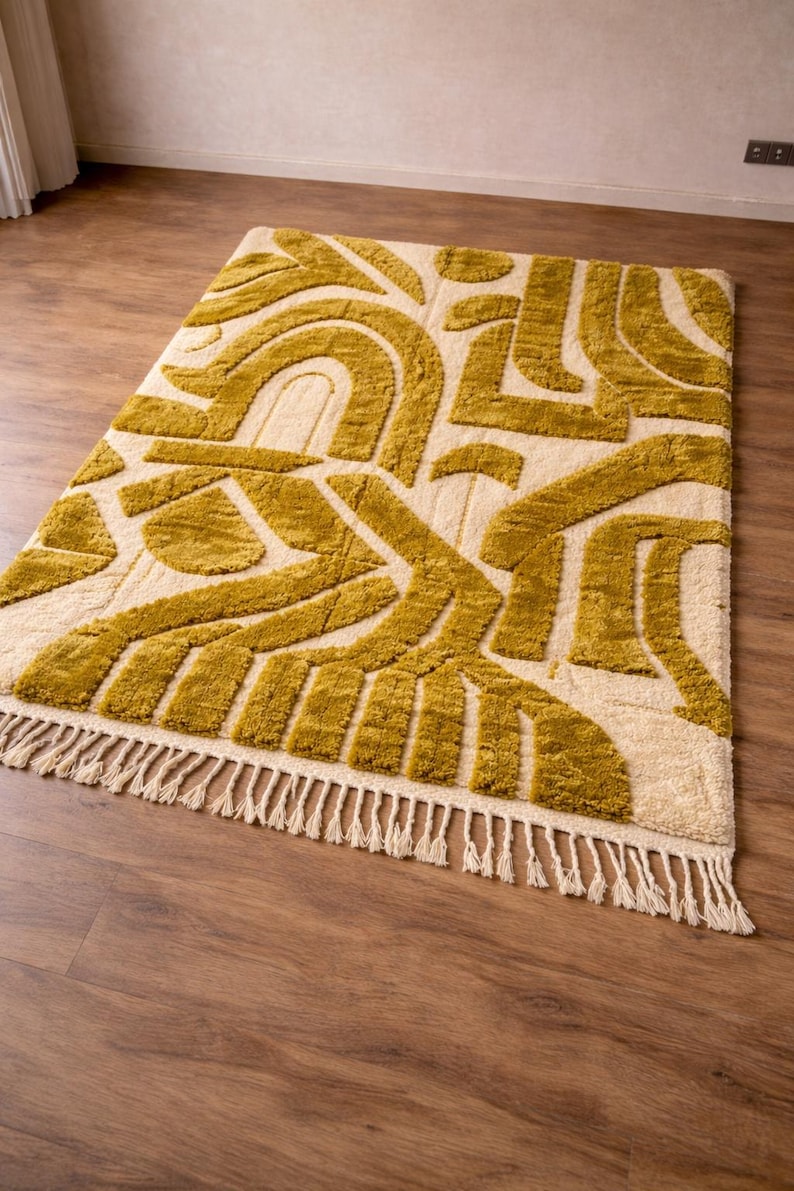 May include: Mustard yellow and cream-colored area rug with an abstract geometric pattern. The rug features a repeating design of curved and angular shapes. The rug has a fringe along one edge. The rug is placed on a hardwood floor.