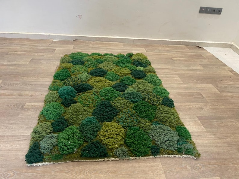 May include: A rectangular rug with a textured surface, featuring a pattern of varying shades of green, resembling a mossy landscape. The rug is placed on a light brown wooden floor.