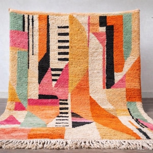 May include: A colourful geometric rug with a cream fringe. The rug features a vibrant design with orange, pink, black, and green shapes. The rug's design is abstract and modern, suitable for home decor.