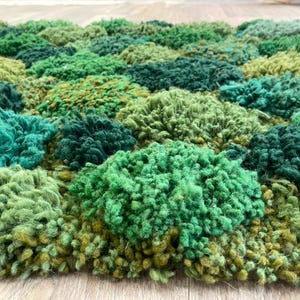 May include: A close-up of a textured rug with a pattern of various shades of green. The rug's surface is made of looped yarn, creating a dense, plush appearance. The colour palette includes light, medium, and dark greens, with some yellow-green accents.
