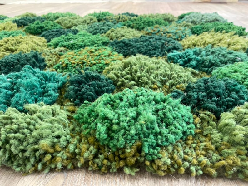 May include: A close-up of a textured rug with a pattern of various shades of green. The rug's surface is made of looped yarn, creating a dense, plush appearance. The colour palette includes light, medium, and dark greens, with some yellow-green accents.