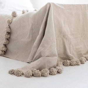 May include: A beige throw blanket with a fringe of fluffy pom-poms. The blanket is draped over a bed and the pom-poms are visible along the bottom edge.