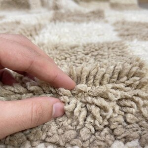 May include: Close-up of a beige and white checkered rug. The rug has a thick, textured pile, with a hand touching the surface. The pattern is a repeating grid of beige and white squares, creating a soft, inviting look.