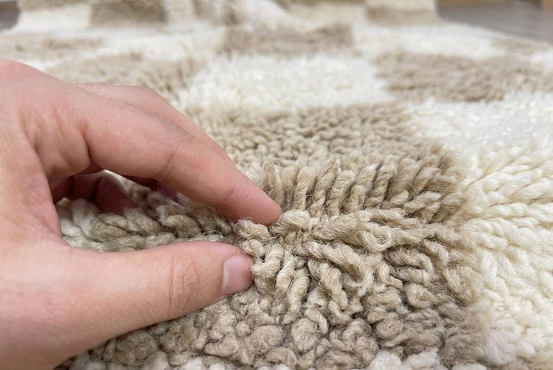 May include: Close-up of a beige and white checkered rug. The rug has a thick, textured pile, with a hand touching the surface. The pattern is a repeating grid of beige and white squares, creating a soft, inviting look.