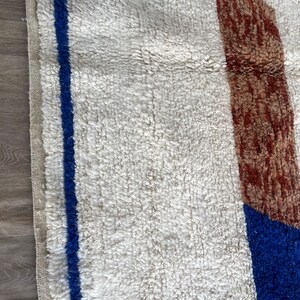 May include: A white rug with a blue and brown geometric pattern. The rug has a thick, fluffy texture.