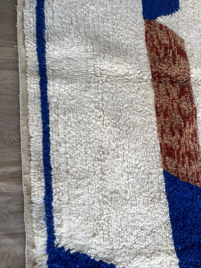 May include: A white rug with a blue and brown geometric pattern. The rug has a thick, fluffy texture.