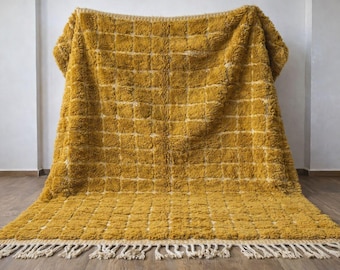 May include: Mustard yellow rug with a grid pattern and a fringed edge. The rug is made of a thick, textured material and is displayed against a white wall. The rug's design features a series of squares, creating a geometric pattern.