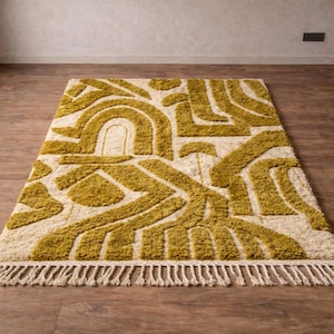 May include: A rectangular rug with a cream-colored background and a geometric pattern in shades of olive green. The design features curved and angular shapes. The rug has a fringe along one edge, adding texture and detail to the floor covering.