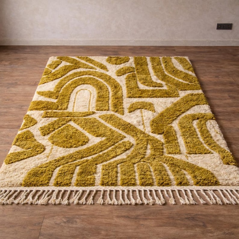 May include: A rectangular rug with a cream-colored background and a geometric pattern in shades of olive green. The design features curved and angular shapes. The rug has a fringe along one edge, adding texture and detail to the floor covering.