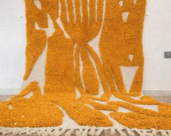 May include: Mustard yellow and off-white tufted rug with an abstract design. The rug features a textured, plush surface with a repeating pattern of stylized shapes and lines. The rug has a fringed edge and is displayed against a white wall.