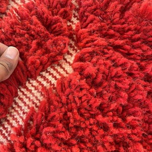 May include: Close-up of a red and cream-colored rug. The rug features a thick, textured pile in a vibrant red hue, with a cream-colored geometric pattern. The design includes a series of cream-colored stripes. The rug's texture is soft and inviting.