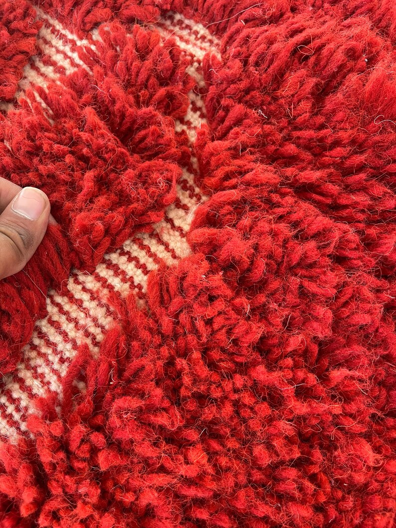 May include: Close-up of a red and cream-colored rug. The rug features a thick, textured pile in a vibrant red hue, with a cream-colored geometric pattern. The design includes a series of cream-colored stripes. The rug's texture is soft and inviting.