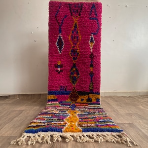 May include: A pink, blue, and yellow Moroccan rug with a geometric pattern. The rug has a fringe border and is made of wool.