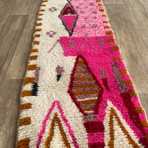 Op de afbeelding: Een lang, rechthoekig tapijt met een witte achtergrond en een kleurrijk geometrisch patroon. Het patroon heeft roze, rode, oranje en bruine kleuren. Het tapijt heeft een franje rand.
