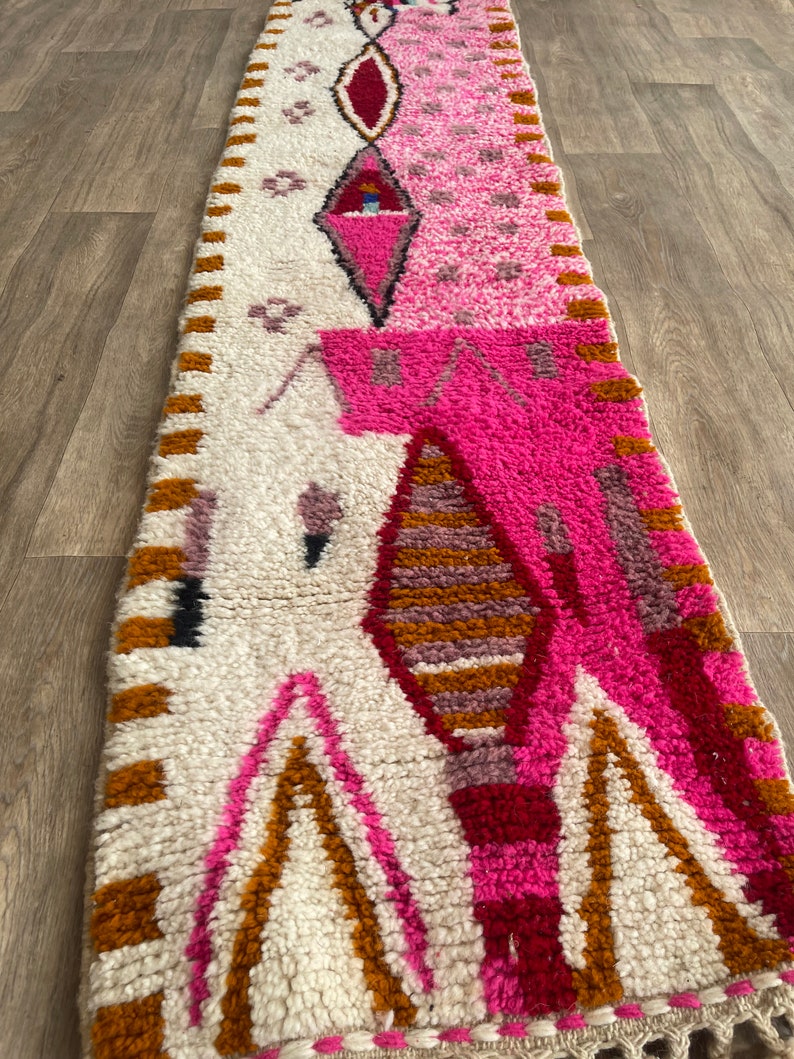 Op de afbeelding: Een lang, rechthoekig tapijt met een witte achtergrond en een kleurrijk geometrisch patroon. Het patroon heeft roze, rode, oranje en bruine kleuren. Het tapijt heeft een franje rand.