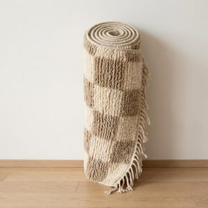 May include: Rolled-up, beige and brown checkered rug with fringe. The rug is made of a textured material and is rolled up against a white wall and wooden floor. The rug's pattern is a series of alternating squares.