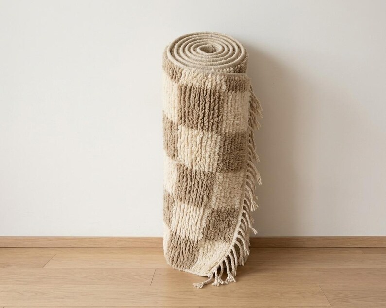 May include: Rolled-up, beige and brown checkered rug with fringe. The rug is made of a textured material and is rolled up against a white wall and wooden floor. The rug's pattern is a series of alternating squares.