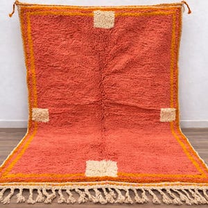 May include: A rectangular, coral-colored rug with a plush texture. It features a border in shades of orange and yellow, with four beige square accents. The rug has a fringe along the bottom edge and is suitable for home decor.