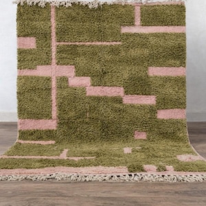 Puede incluir: Una alfombra rectangular y peluda con un diseño geométrico. La alfombra presenta un color base verde oliva con un patrón de rectángulos y líneas rosas. Los bordes están acabados con flecos.
