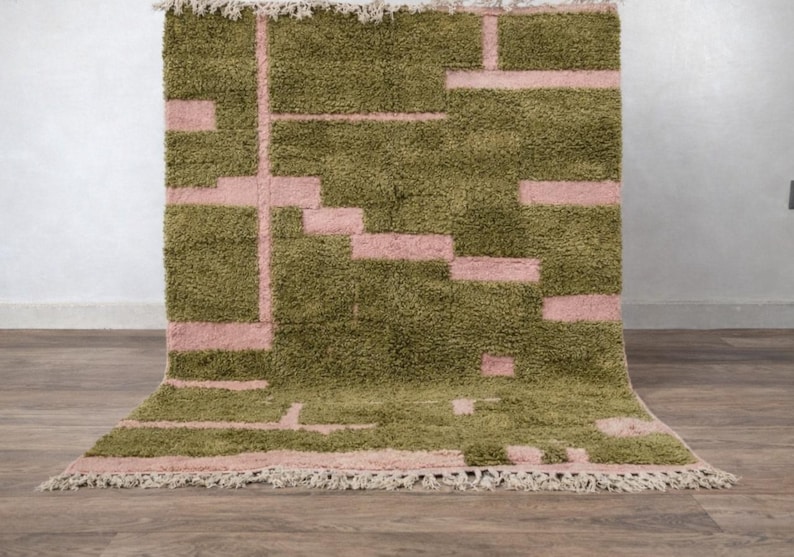Puede incluir: Una alfombra rectangular y peluda con un dise&ntilde;o geom&eacute;trico. La alfombra presenta un color base verde oliva con un patr&oacute;n de rect&aacute;ngulos y l&iacute;neas rosas. Los bordes est&aacute;n acabados con flecos.