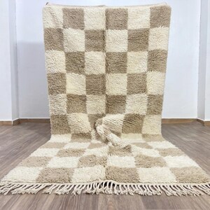 May include: A large, rectangular rug with a checkerboard pattern in cream and tan. The rug has a thick, textured pile and a fringe along the bottom edge. The rug is displayed on a wooden floor.