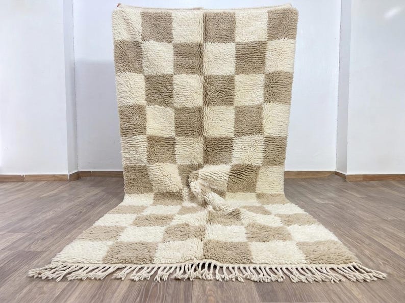 May include: A large, rectangular rug with a checkerboard pattern in cream and tan. The rug has a thick, textured pile and a fringe along the bottom edge. The rug is displayed on a wooden floor.