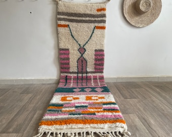 May include: A long, rectangular rug with a white background and a geometric pattern in shades of pink, orange, green, and grey. The rug has a fringe on the bottom.