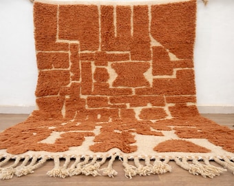 May include: A rectangular, hand-woven rug with a geometric pattern in shades of orange and cream. The rug features a textured, raised design and a fringe along the bottom edge. The rug's design includes rectangular and abstract shapes. This rug is suitable for living rooms and bedrooms.