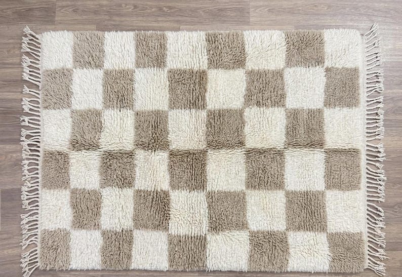 May include: A rectangular, checkered rug with a neutral color palette. The rug features a pattern of alternating beige and off-white squares. The rug has a shaggy texture and fringe detailing along the edges. The rug is designed for home decor.