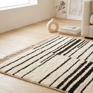 May include: A rectangular area rug featuring a cream-colored base with a pattern of dark brown horizontal stripes of varying widths. The rug has a textured, shaggy pile and is finished with short, knotted fringe along the shorter edges.