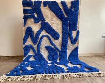 May include: A rectangular rug with a cream-colored background and bold blue abstract shapes. The rug has a textured, plush appearance, with a fringe along the bottom edge. The design is modern and eye-catching.