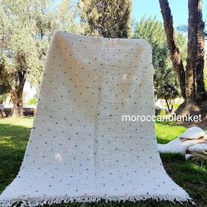 Puede incluir: Alfombra marroquí blanca con borde de flecos y puntos de colores esparcidos por toda la alfombra. La alfombra se muestra en un césped.