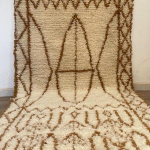 May include: A cream-coloured, textured rug with a geometric pattern in shades of brown. The rug features a central design of diamond and oval shapes, with a border of zigzags and other geometric elements. The rug is made of a soft, plush material.