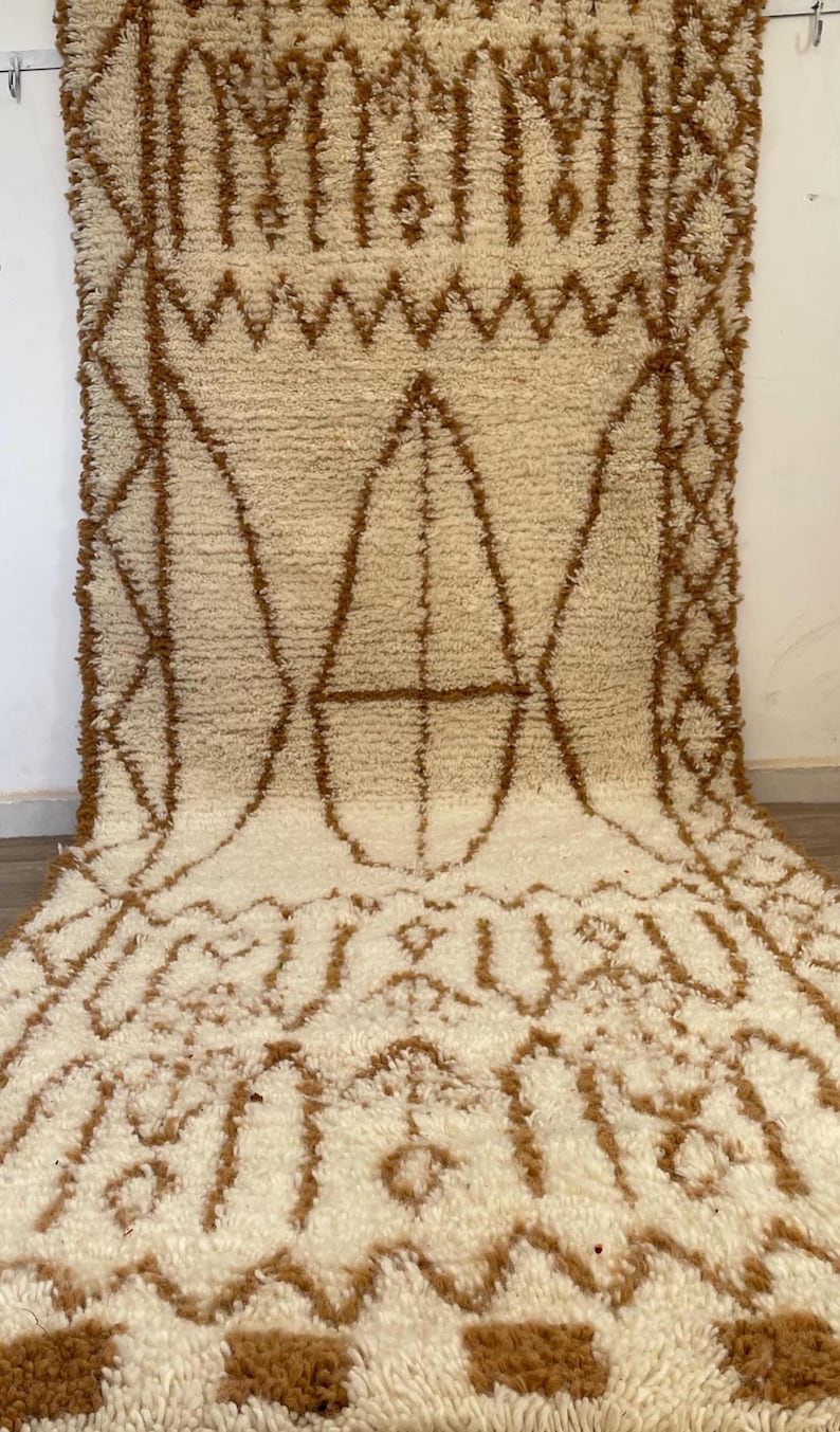 May include: A cream-coloured, textured rug with a geometric pattern in shades of brown. The rug features a central design of diamond and oval shapes, with a border of zigzags and other geometric elements. The rug is made of a soft, plush material.
