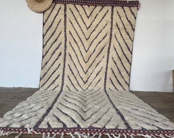 Puede incluir: Una alfombra marroquí blanca con un patrón de chevron en tonos de morado y azul. La alfombra tiene un borde de flecos y está hecha de lana.