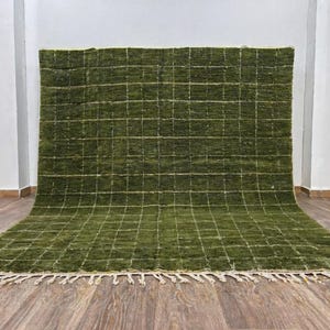 May include: A large, rectangular green rug with a grid pattern. The rug has a thick pile and a fringe along the bottom edge. The grid lines are a lighter shade of green. The rug is displayed on a wooden floor.