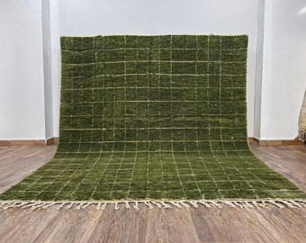 May include: A large, rectangular green rug with a grid pattern. The rug has a thick pile and a fringe along the bottom edge. The grid lines are a lighter shade of green. The rug is displayed on a wooden floor.
