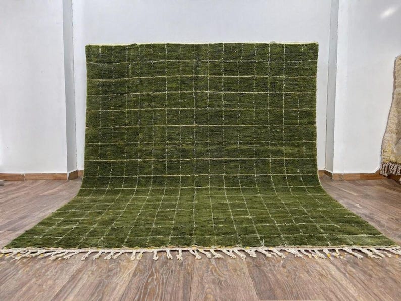 May include: A large, rectangular green rug with a grid pattern. The rug has a thick pile and a fringe along the bottom edge. The grid lines are a lighter shade of green. The rug is displayed on a wooden floor.